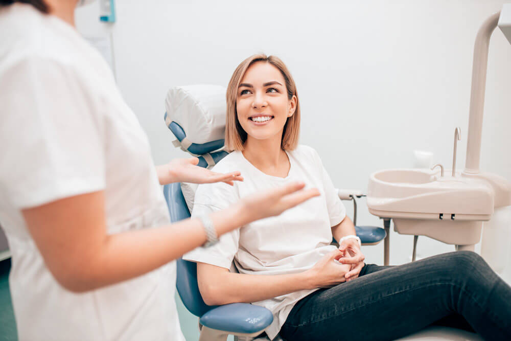 Blonde woman sitting in dentist office char speaking with a dentist 